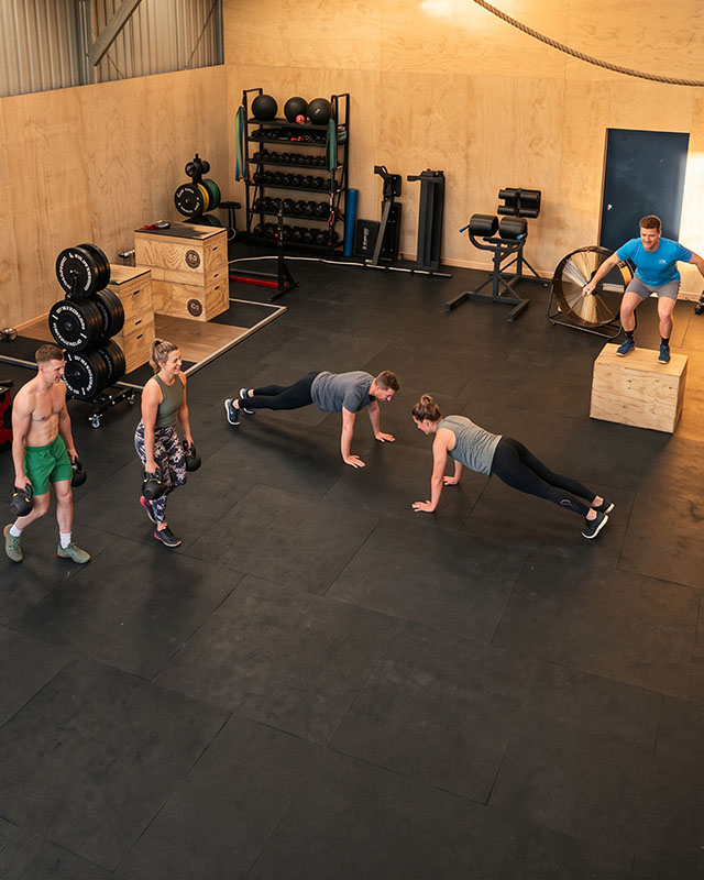 Small group of four athletes training together in an industrial gym with a coach guiding the session, sense of camaraderie, wide shot showing lifting platforms and rig in the background, warm golden hour lighting