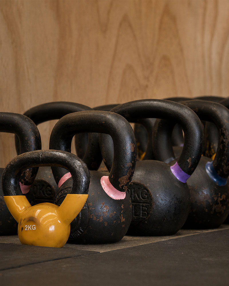 Detail shot of strength training equipment lined up against a wall — kettlebells, dumbbells and wall balls arranged by size, chalk dust visible, warm directional light, editorial still-life fitness photography