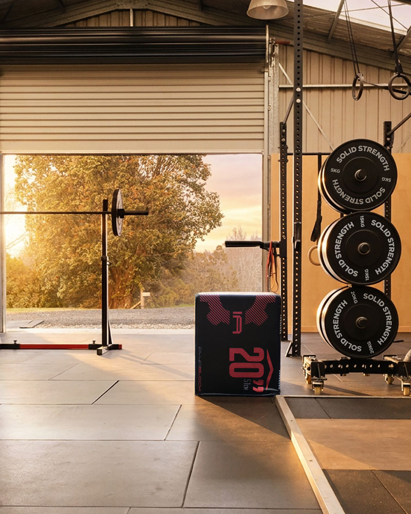 Athletes training outdoors just beyond an open roller door, using kettlebells and wall balls on a concrete apron, rural New Zealand paddocks visible in the background, natural daylight, editorial fitness photography